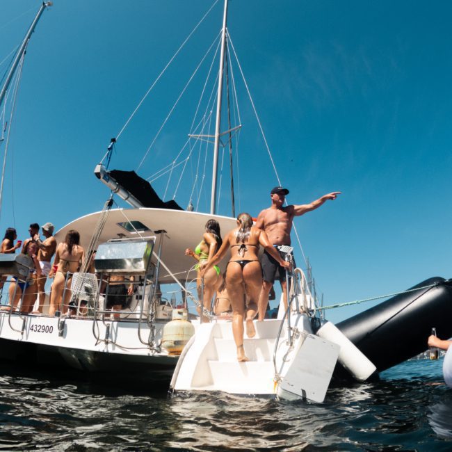 People in swimwear are gathered on a white boat in the water. One person gestures while others socialize, enjoying the Sydney boat party hire. Another boat and inflatable rafts are nearby under a clear blue sky.