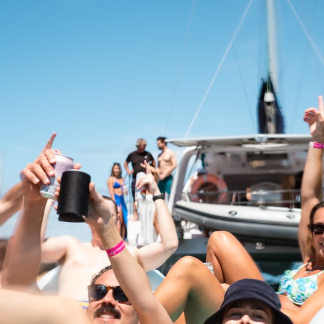Group of people in swimwear enjoying a catamaran party on Sydney Harbour, some holding drinks and raising their hands, with a luxury yacht in the background under a clear blue sky.
