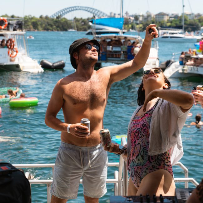Three people stand on a boat deck, holding drinks and enjoying a sunny day. Inflatable toys float in the water, with the Sydney Harbour Bridge visible in the background, setting the perfect scene for a catamaran party Sydney style.