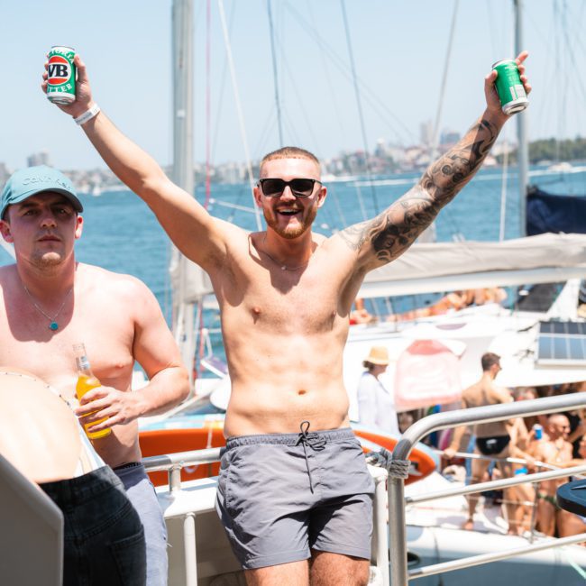 A shirtless man with sunglasses raises two cans of beer aboard a luxury yacht, surrounded by other people.