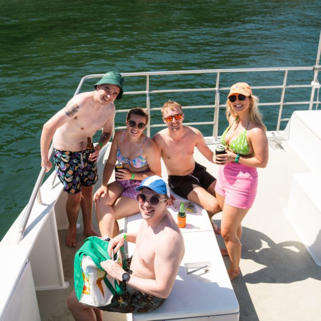 A group of five people wearing summer clothing are standing and sitting on a boat, smiling and holding drinks. The catamaran party Sydney is on a body of water, and it appears to be a sunny day.