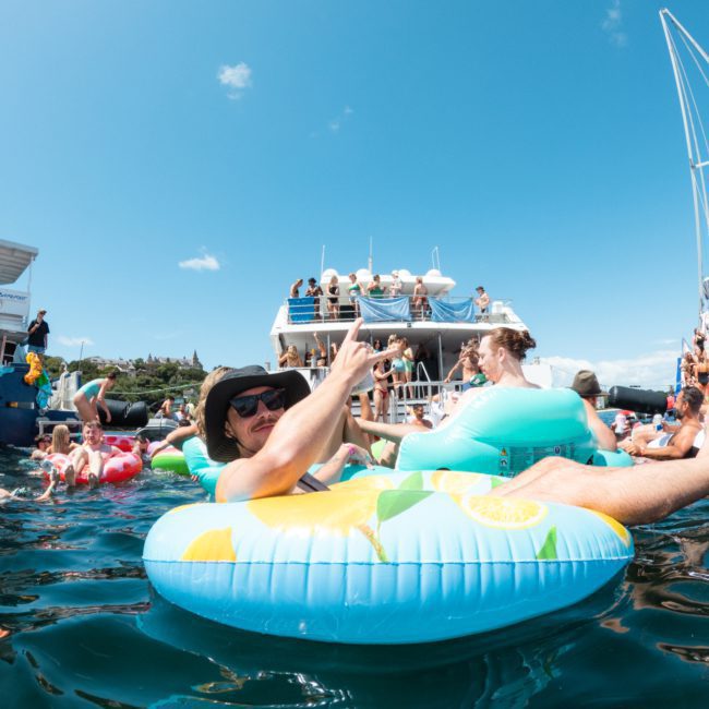 People are enjoying a sunny day on the water, floating on inflatable rings and socializing near boats, with some individuals diving into the water. It's the perfect scene for a luxury yacht hire Sydney experience.