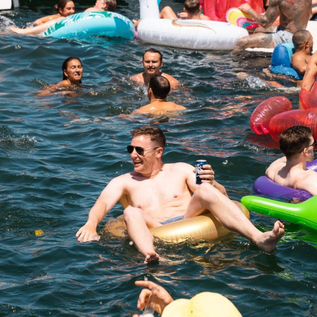 A group of people enjoying a sunny day in the water, floating on inflatable toys and holding drinks. Some are swimming near a Sydney boat party hire, while a luxurious yacht is visible in the background.