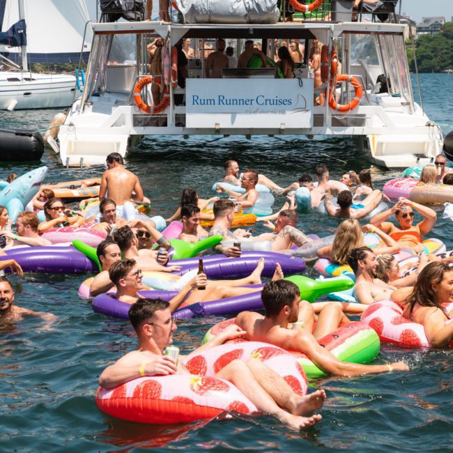 A group of people relax on inflatable floats in the water near a boat labeled "Rum Runner Cruises." The boat is docked, and the scene is lively with many enjoying the sun and drinks, embodying the perfect vibe for Sydney boat party hire.