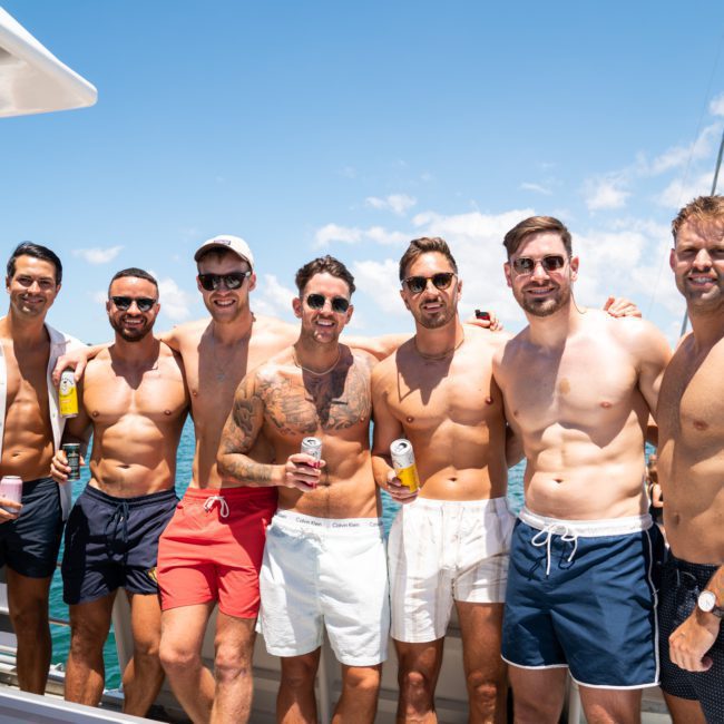 A group of seven men in swimwear standing on a boat, holding drinks, with clear blue sky and water in the background. Perfect for those looking to experience a Sydney boat party hire with friends or colleagues.