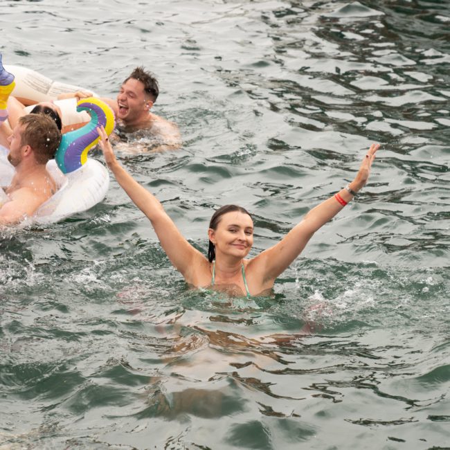 A woman in a green swimsuit swims with her arms raised among other people in an outdoor water setting with inflatable toys, including a unicorn. The scene looks like the fun you'd find at a Sydney boat party hire.