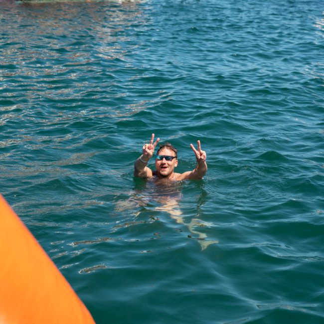 A man is swimming in the water, raising both hands to make peace signs, with a large group of people standing on a dock in the background, likely enjoying a lively Sydney boat party hire.