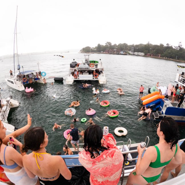 A group of people enjoy a party on a luxury yacht hire Sydney, surrounded by inflatable pool floats near the shoreline. Some are dancing and socializing, while others swim or float in the water.