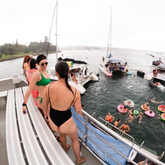 A group of people in swimsuits are seen socializing on a private yacht charter in Sydney Harbour on a cloudy day. Some are standing on the deck, while others are floating on colorful inflatable rings near other boats.