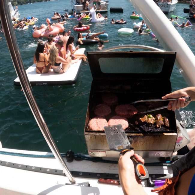 A person grills hamburgers on a boat while people relax on floats and swim in a lake. With other boats and people visible in the background, it sets the perfect scene for a Corporate boat event in Sydney.