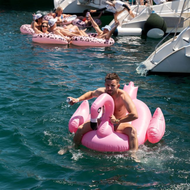 A man rides a pink flamingo float while holding a beverage in a large body of water near several yachts. Other people are also seen relaxing on floats and on the boats, enjoying the sun. Experience this vibe with a private yacht charter Sydney Harbour or DJ boat hire Sydney for your next event!