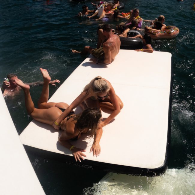 Several people are enjoying a sunny day on the water, some on a large floating platform and others in colorful inflatable tubes. Two women are playfully interacting on the platform as a DJ boat hire Sydney enhances the lively atmosphere.