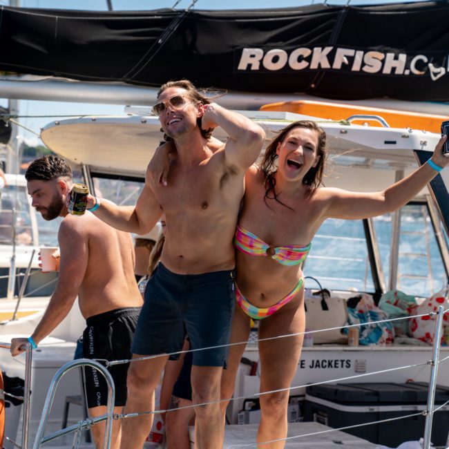 Three people in swimwear are on a boat, holding drinks and smiling. The boat has "ROCKFISH.C....MAUI" written on the canopy. Another person sits nearby, occupied with something, enjoying what looks like a perfect day for a Sydney boat party hire.