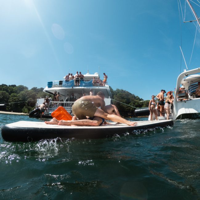 People swimming, relaxing on a paddleboard, and socializing near several boats in the water on a sunny day. It's the perfect setting for a Sydney boat party hire or enjoying a private yacht charter in Sydney Harbour.