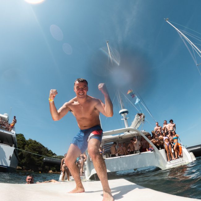 A man poses enthusiastically on a boat with a group of people in the background, enjoying a sunny day on the water at a vibrant Sydney boat party hire.