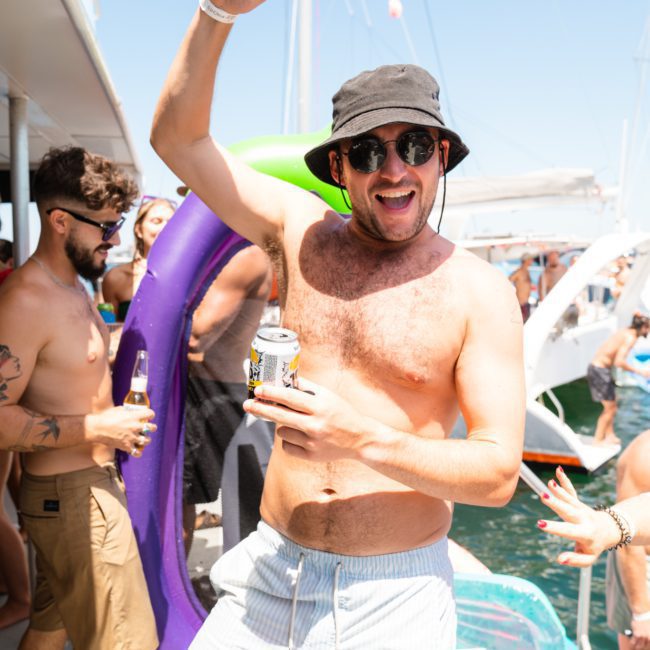 A man wearing sunglasses and a black hat smiles and raises his arm while holding a can of beer, standing on a private yacht charter at a lively party with other people in the background.