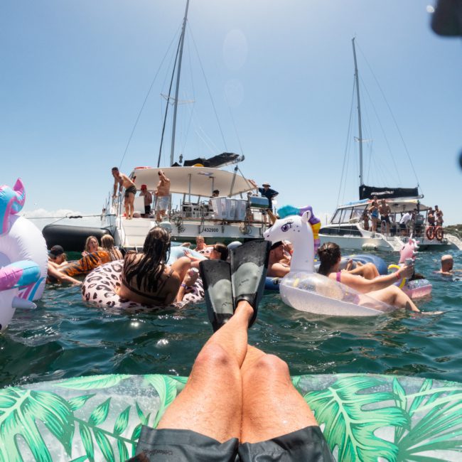 View from someone's perspective relaxing on an inflatable raft with flippers on their feet, surrounded by other people on floats and boats in the background on a sunny day at a Sydney boat party hire.