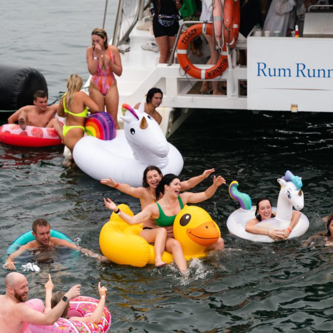 A group of people in swimsuits float on inflatable toys, including a unicorn and a duck, near a catamaran party Sydney boat labeled "Rum Runner Cruises." Some are in the water, and others are about to jump in.