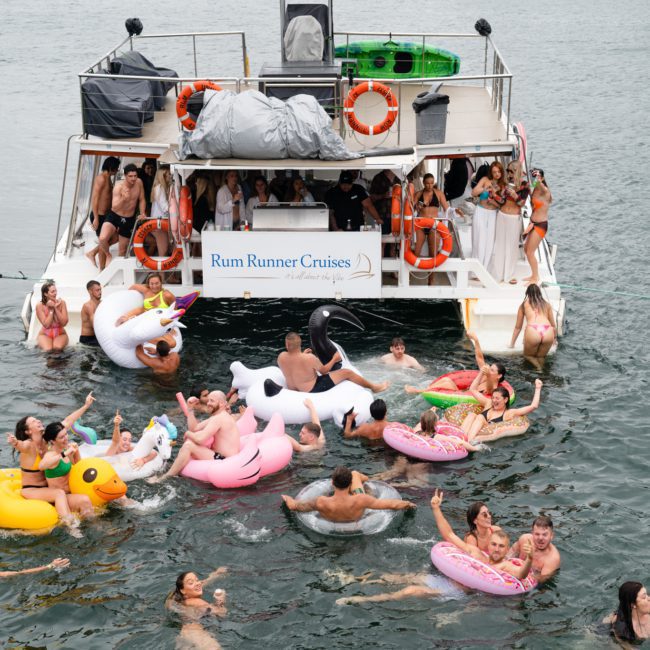 A group of people enjoying a party on a boat and in the water with various inflatable floats, including ducks and swans, near a boat with a "Rum Runner Cruises" banner. This Luxury yacht hire Sydney experience epitomizes fun and relaxation on the water.