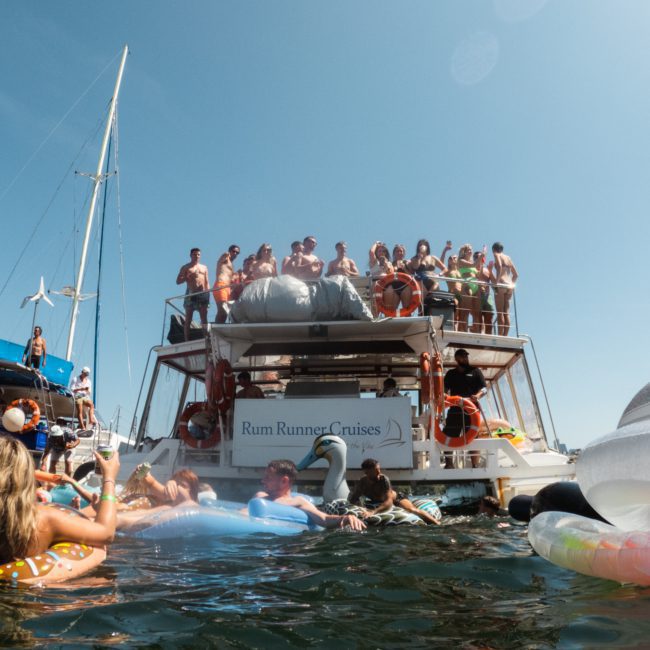 A group of people are on a boat and in the water, enjoying a sunny day. The boat, named Rum Runner Cruises, is crowded with people in swimwear. Several floatation devices are visible in the water, making it look like the perfect Sydney boat party hire event.