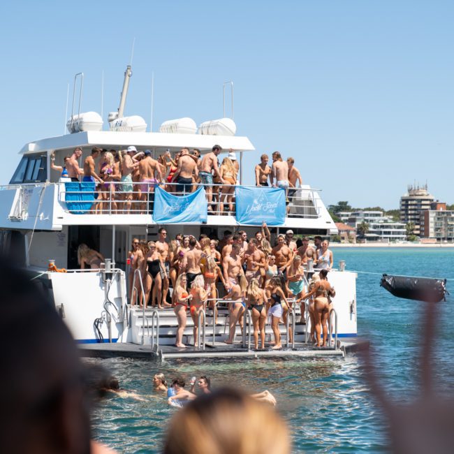 A large group of people in swimwear gather on the deck of a white boat, some swimming in the water, with buildings visible in the background, enjoying a Luxury yacht hire Sydney experience.