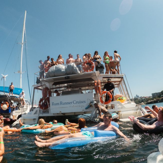 People are enjoying a vibrant Catamaran party Sydney on a vessel labeled "Rum Runner Cruises," with some swimming and lounging on floating devices in the surrounding water.