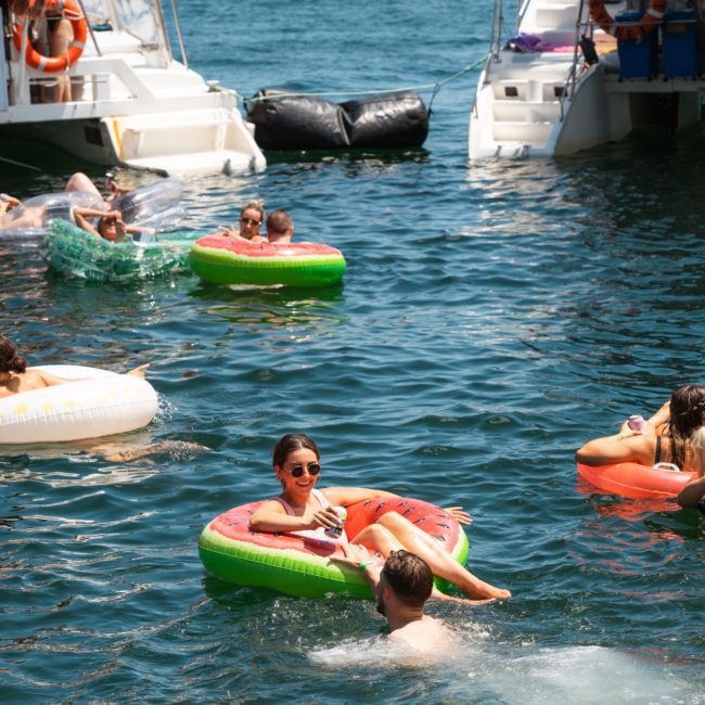 People enjoying a sunny day on the water, some on inflatable pool floats near boats. A woman is lounging on a watermelon float, holding a drink, as others revel in the atmosphere of their private yacht charter on Sydney Harbour.