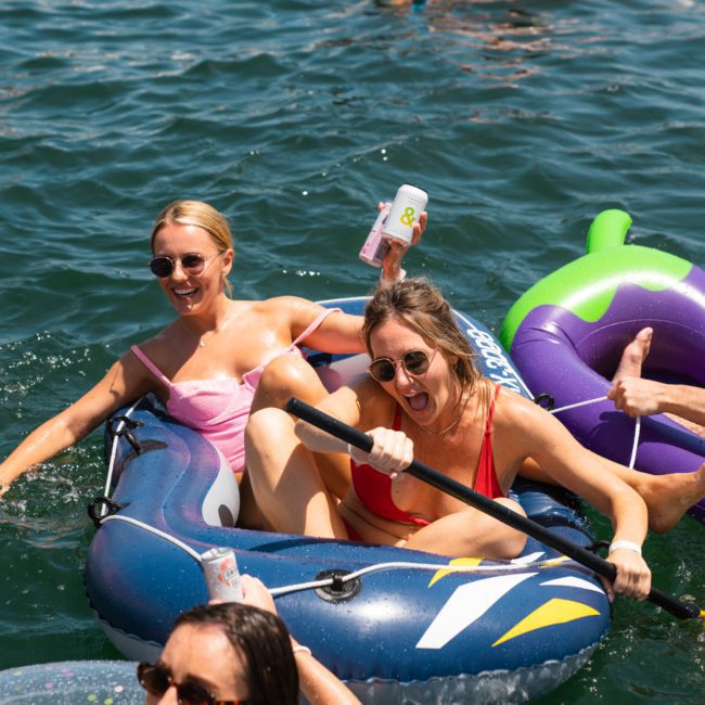 People in swimsuits are enjoying a sunny day on the water, floating on inflatable rafts and inner tubes. Two women in the foreground are paddling an inflatable boat, while nearby a luxury yacht hire Sydney sails in the background.
