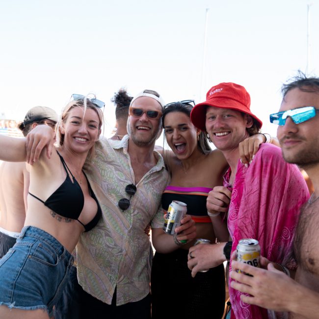 Five people smiling and posing together outdoors on a luxury yacht hire Sydney. They are dressed in casual summer clothing, some holding beverages, and appear to be enjoying a private yacht charter Sydney Harbour gathering or party near water.
