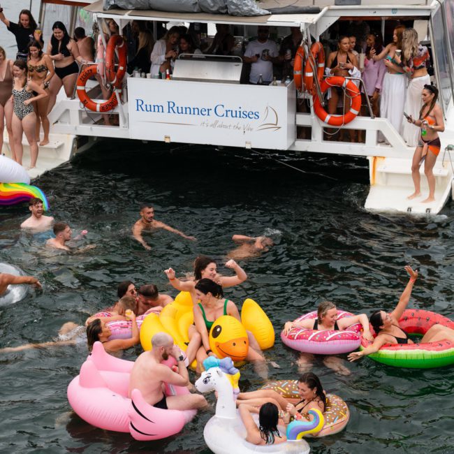 A group of people enjoys a Catamaran party Sydney on the water, with many floating on various inflatable rafts, near a boat labeled "Rum Runner Cruises.
