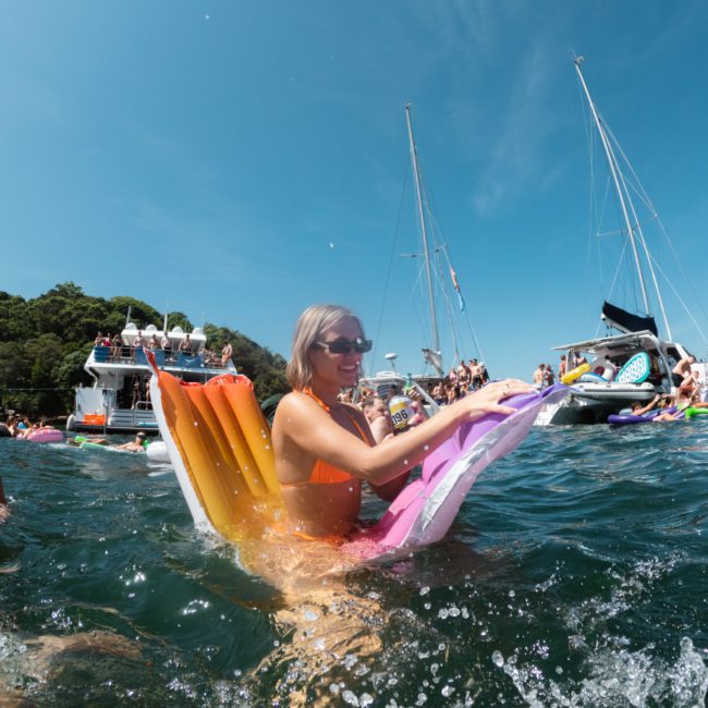 A person in a bikini lounges on a colorful inflatable in the water, holding a beverage, with boats and other people enjoying themselves in the background under a clear blue sky. Consider booking your own private yacht charter on Sydney Harbour for an unforgettable experience.