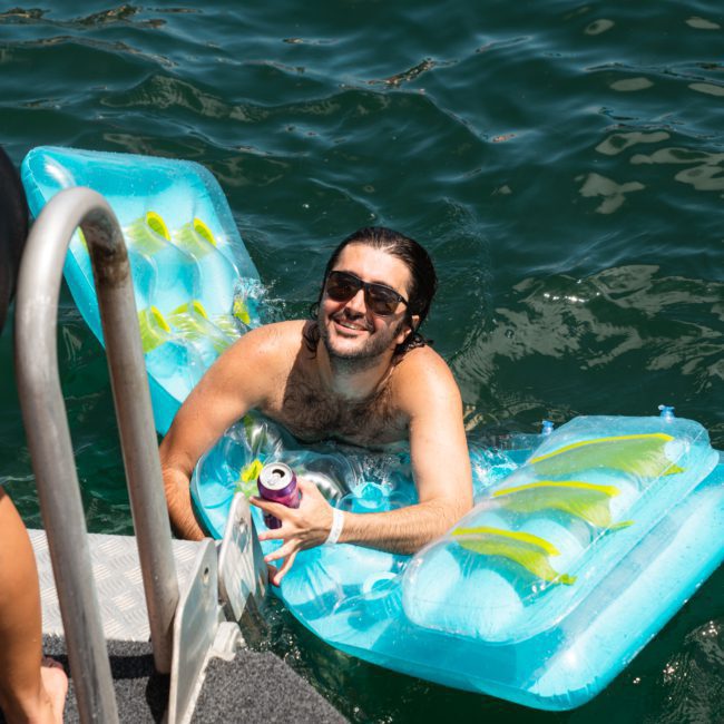 A man with long hair and sunglasses relaxes on an inflatable float in a body of water, holding a canned beverage. Part of a metal ladder and a person’s leg are visible on the left, hinting at the luxury yacht hire Sydney experience.