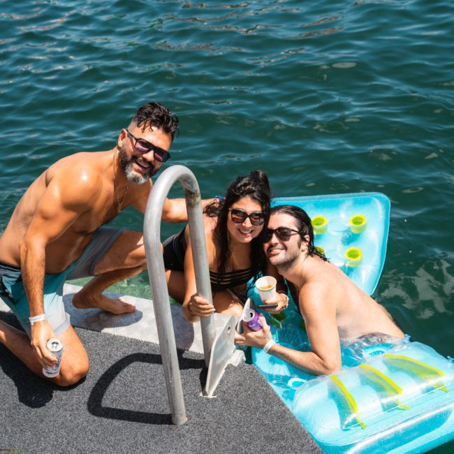 Three people, two standing on a dock and one seated on an inflatable float in the water, smiling and posing for a photo. They appear to be enjoying a sunny day at the lake, reminiscent of a lively Catamaran party Sydney has to offer.
