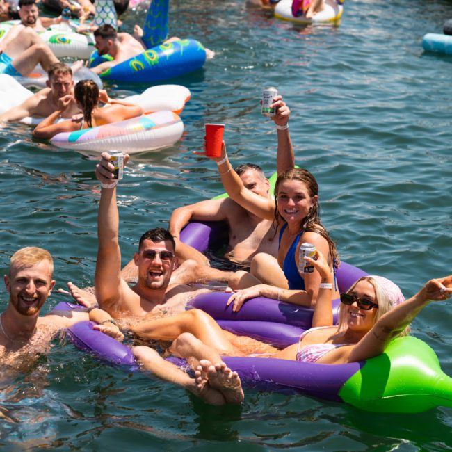 A group of people floating in an inflatable on water, smiling and raising their drinks during a Catamaran party in Sydney. More individuals can be seen on various inflatables in the background.