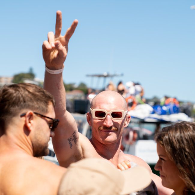 A bald man with sunglasses is raising his hand and making a peace sign gesture in a crowd of people at a private yacht charter on Sydney Harbour.