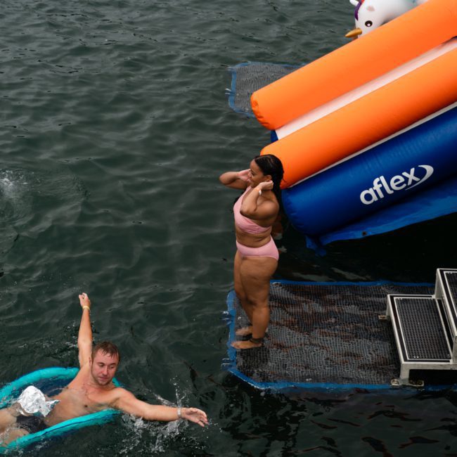 People are swimming and using inflatable floats in a body of water near an aflex slide. A woman in a pink bathing suit stands on a platform next to the slide, while a DJ boat hire Sydney creates lively music for the atmosphere.