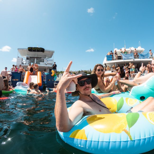 People are floating in inflatable tubes in the water near a catamaran party on a sunny day. One person in the foreground is making a peace sign.