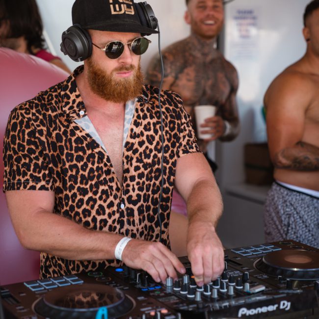 A bearded DJ wearing a leopard print shirt and sunglasses mixes music on a boat during a luxury yacht hire in Sydney Harbour. Other people are in the background, including one shirtless man with tattoos.