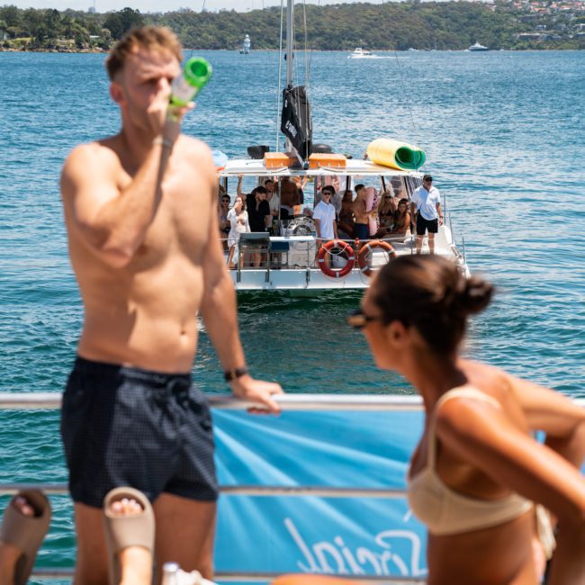 Two people in swimwear are relaxing on a private yacht charter in Sydney Harbour. One is drinking from a green bottle while another boat full of people sails by on this sunny day.