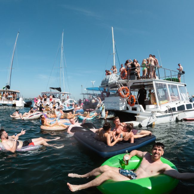 People are enjoying a sunny day on the water, relaxing on inflatables and boats. Some are socializing, sunbathing, or swimming near a group of anchored boats on a clear day, creating the perfect scene for a luxury yacht hire Sydney event.