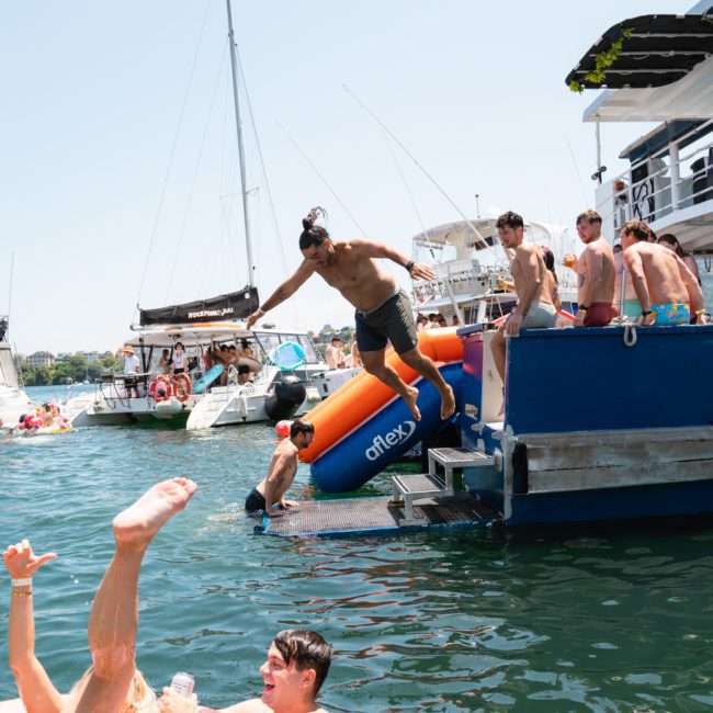 A person jumps into the water from a boat, while others watch. Nearby people are floating in inflatable rings. Several boats, possibly from a Sydney boat party hire, are docked in the background on this sunny day.
