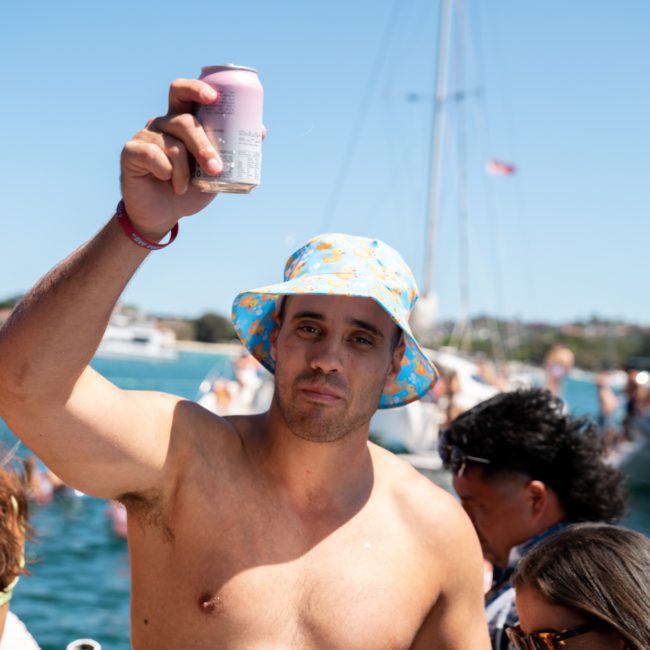 A shirtless man in a blue bucket hat holds up a beverage can on a crowded boat, surrounded by people and other boats in the background, enjoying what seems like a lively Sydney boat party hire.