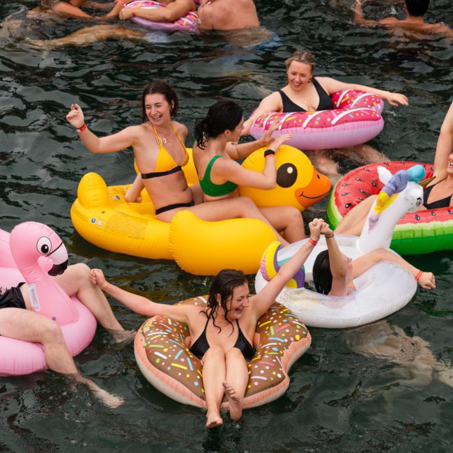 A group of people in swimsuits enjoys floating on inflatable pool toys, including a flamingo, duck, donut, watermelon, and unicorn, in the water while holding hands and cheering during a private yacht charter on Sydney Harbour.