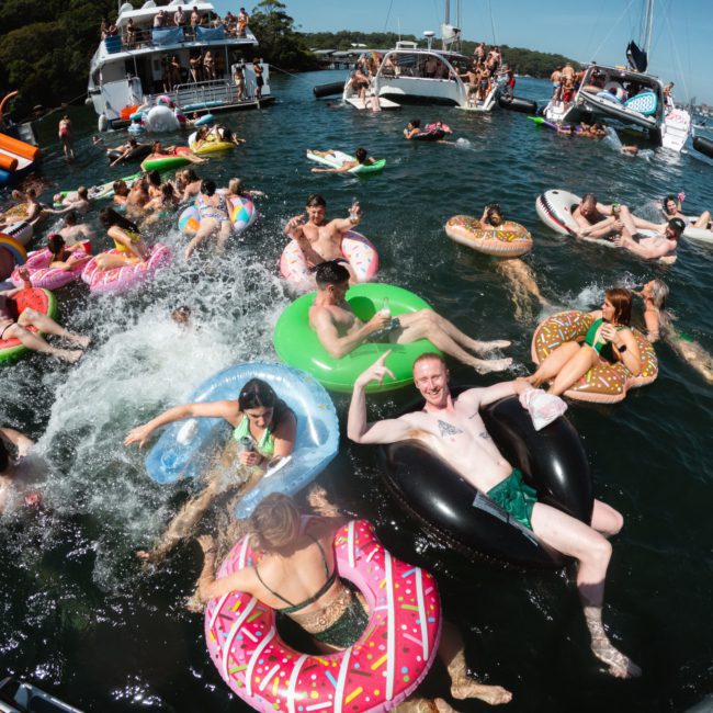 A group of people enjoy a sunny day on the water, floating on inflatable pool toys while boats are anchored nearby. Some are sitting on the floats, while others swim and splash. The scene is enhanced by a private yacht charter in Sydney Harbour and DJ boat hire for lively music.