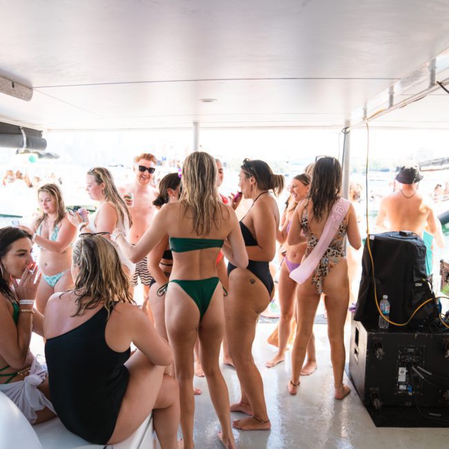A group of people in swimwear socialize on a private yacht charter in Sydney Harbour, with others seen on nearby boats in the background. The scene is lively and appears to be a party.