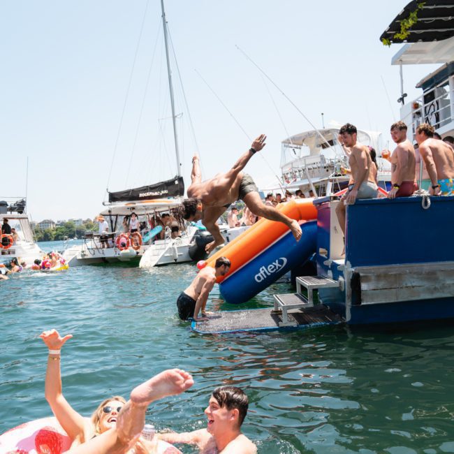 People enjoying a sunny day on boats in the water; some are jumping in, while others are on inflatables. A few are indulging in luxury yacht hires around Sydney Harbour, making the most of their time.