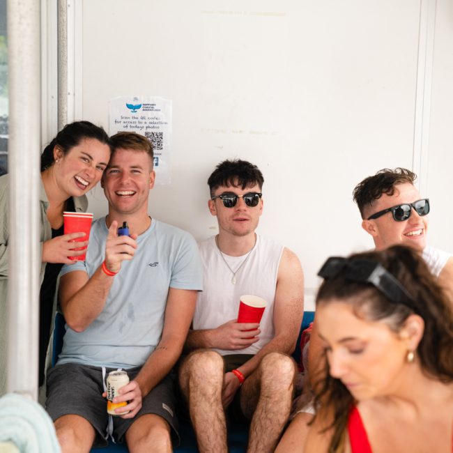 A group of people sits together indoors, smiling and holding red cups. One person stands to the left, and another sits to the far right. They appear to be enjoying a social gathering, reminiscent of a corporate boat event in Sydney Harbour.