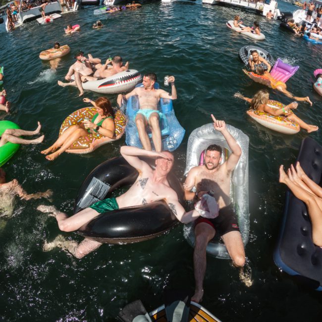 People relaxing on inflatable floaties in the water, surrounded by anchored boats on a sunny day. Some are swimming, while others float and enjoy the scene at a lively Sydney boat party hire event.
