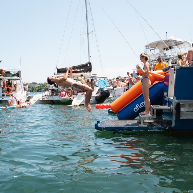 A person backflips off a boat into the water while others watch. Numerous people and boats are in the background, with some people in inflatables. The scene is on a sunny day, perfect for a private yacht charter on Sydney Harbour.