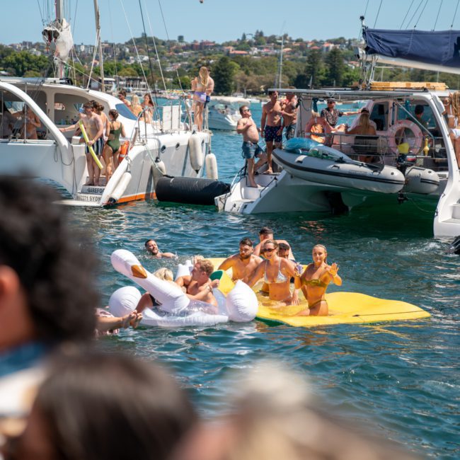 People enjoying a boat party on a lake with some floating on a yellow mat, others on boats, and a person dressed as a unicorn. The weather is sunny, and the background shows houses and trees. It's the perfect scene for DJ boat hire in Sydney.